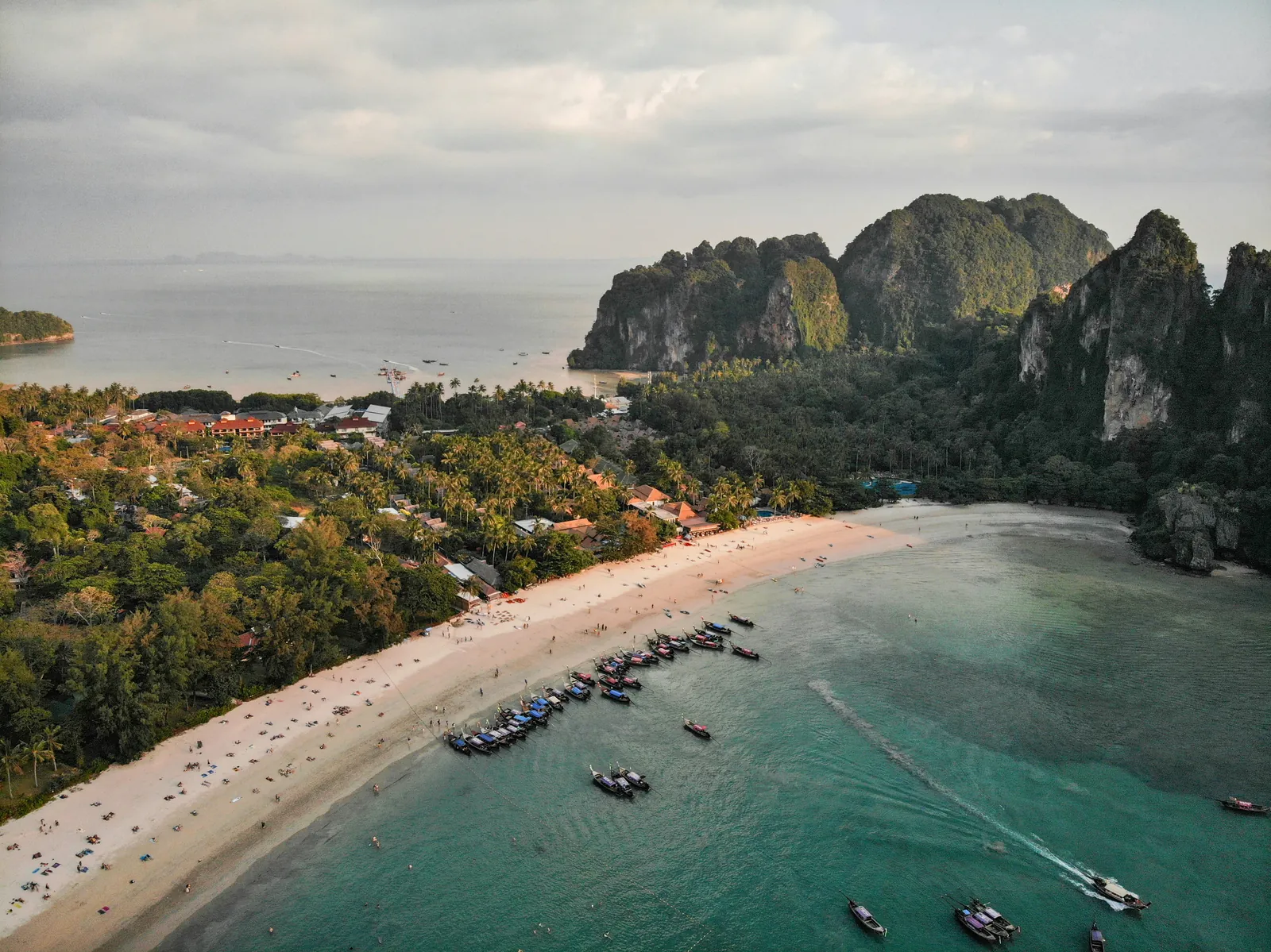 Aerial view of a long pale beach lined with palms, longtail boats in turquoise water and tall limestone cliffs on the right