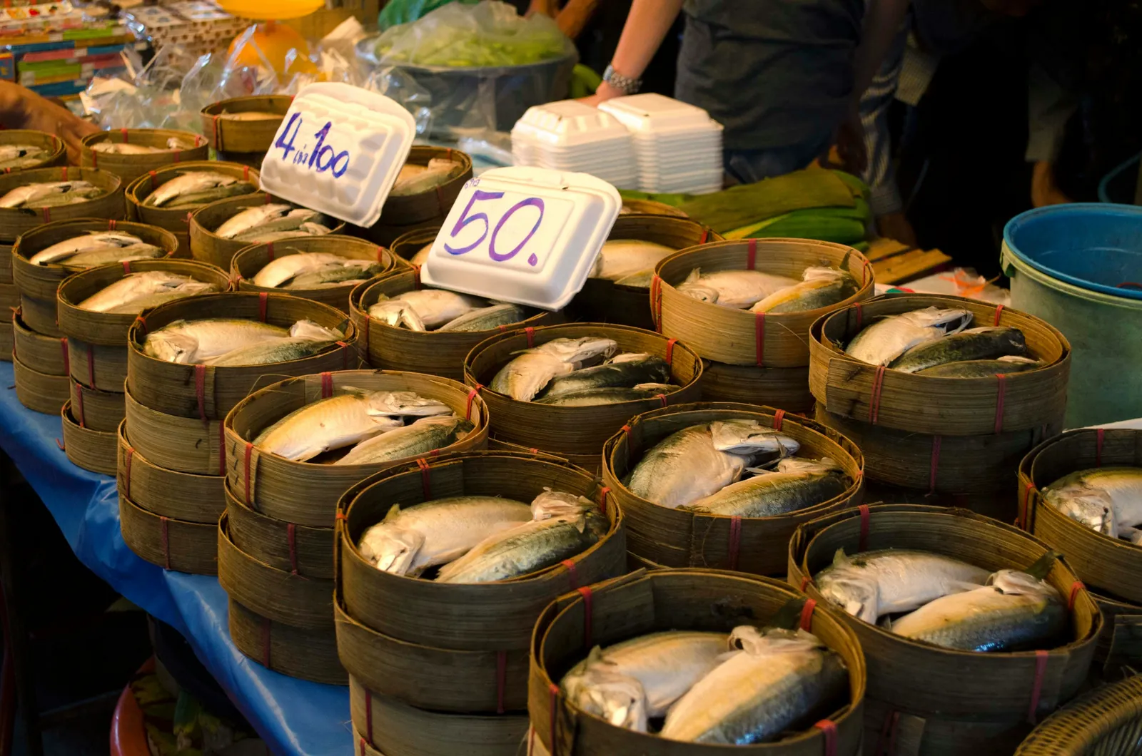 Rows of small bamboo baskets of steamed mackerel on a market table, with handwritten price cards reading 4 for 100 and 50