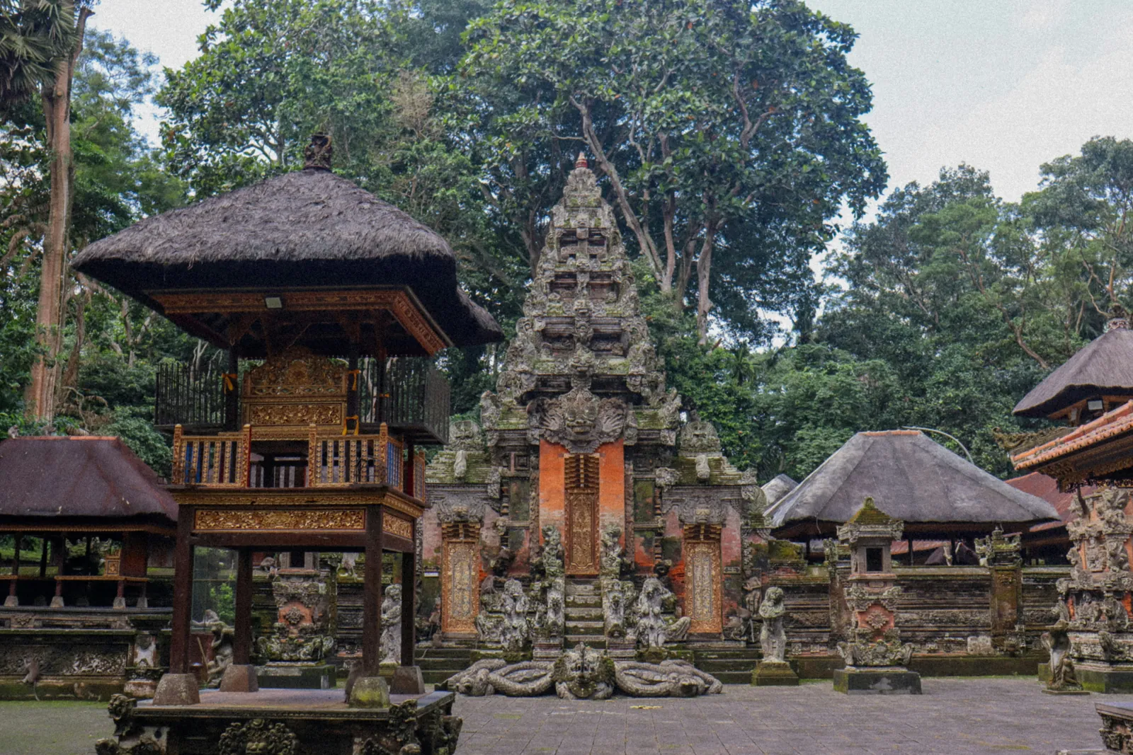 A temple courtyard in the Monkey Forest, with a carved stone gateway, a raised pavilion with a thatched roof and stone statues under tall trees
