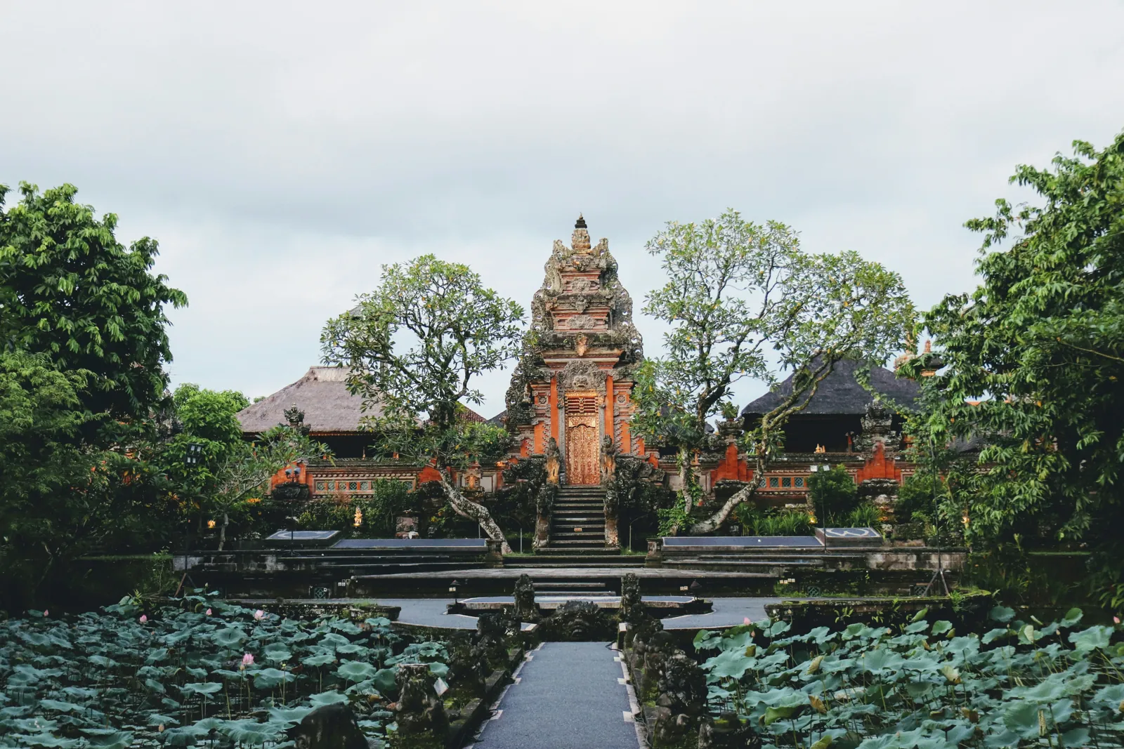 A carved temple gateway at the end of a stone path through a lotus pond, with old trees on both sides and a grey sky