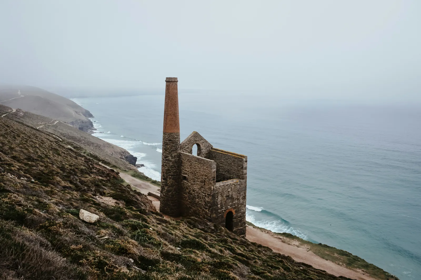 Ruined stone engine house and tall brick-topped chimney on a heather slope above the sea, with surf and cliffs fading into mist
