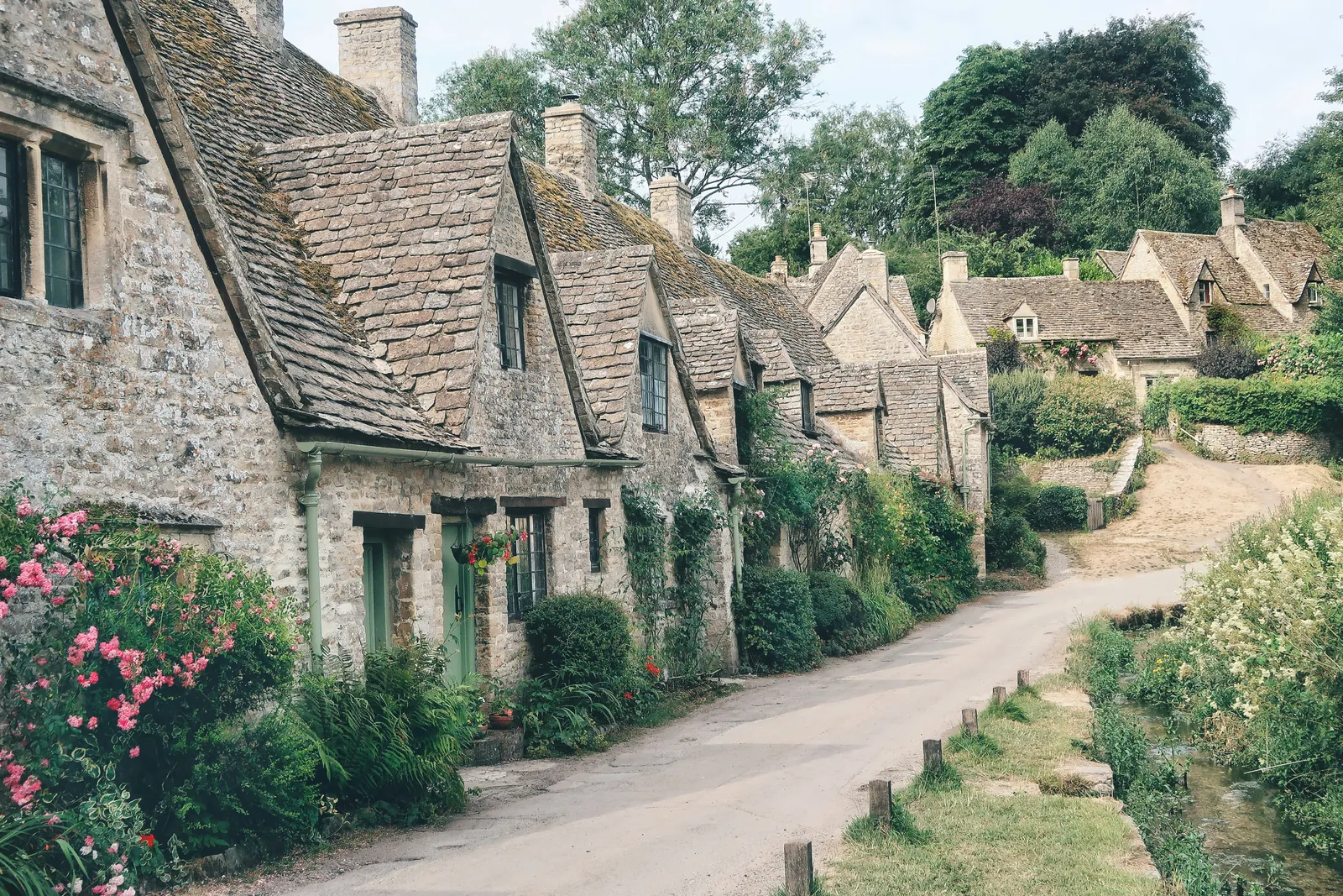 A row of old honey-coloured stone cottages with steep stone-tiled roofs and green doors, roses in front and a lane curving uphill past a small stream