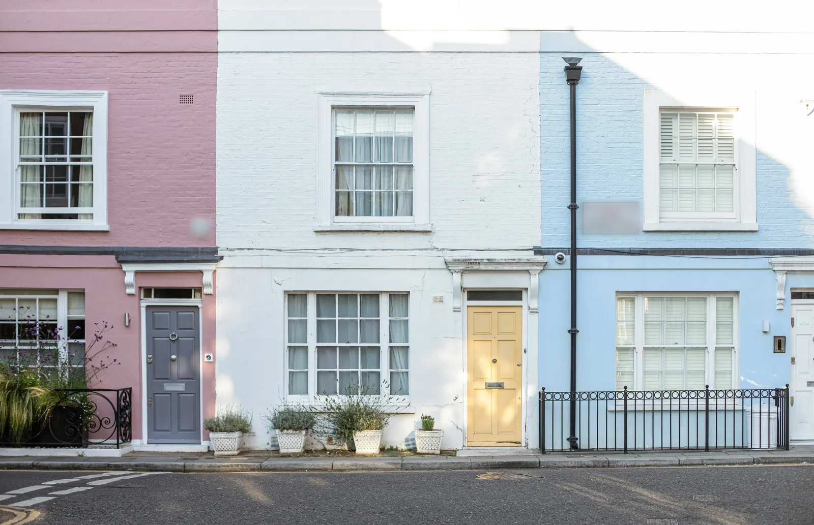 Three small terraced houses painted pink, white and pale blue, with sash windows, a grey door and a yellow door, pots of lavender and black railings on a quiet street
