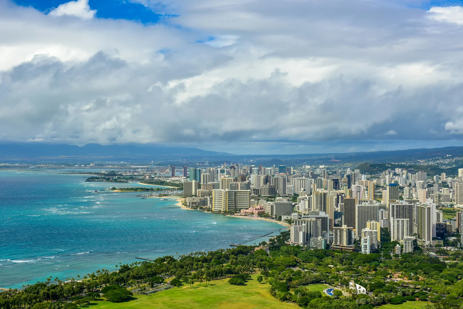 A view from high above a green park over a curving shoreline of hotel and apartment towers beside turquoise water, with mountains under heavy white cloud behind