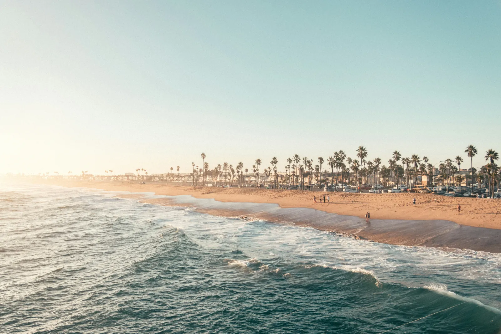 Low golden sun over a wide sandy beach with waves rolling in, a row of tall palm trees and beach houses behind, and a few small figures on the sand