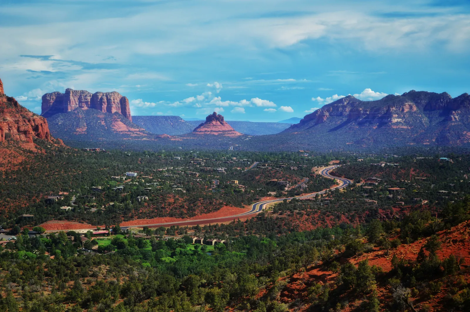 A winding road through a green valley of houses and juniper trees, ringed by red sandstone buttes and mesas under a blue sky