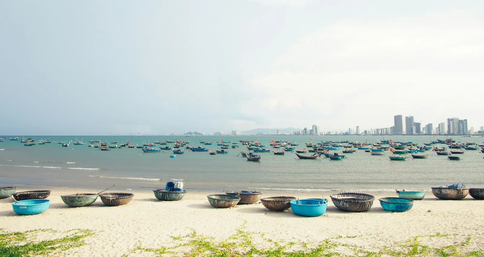 Round basket boats pulled up on white sand, with fishing boats moored in the bay and a line of high-rise towers on the far shore