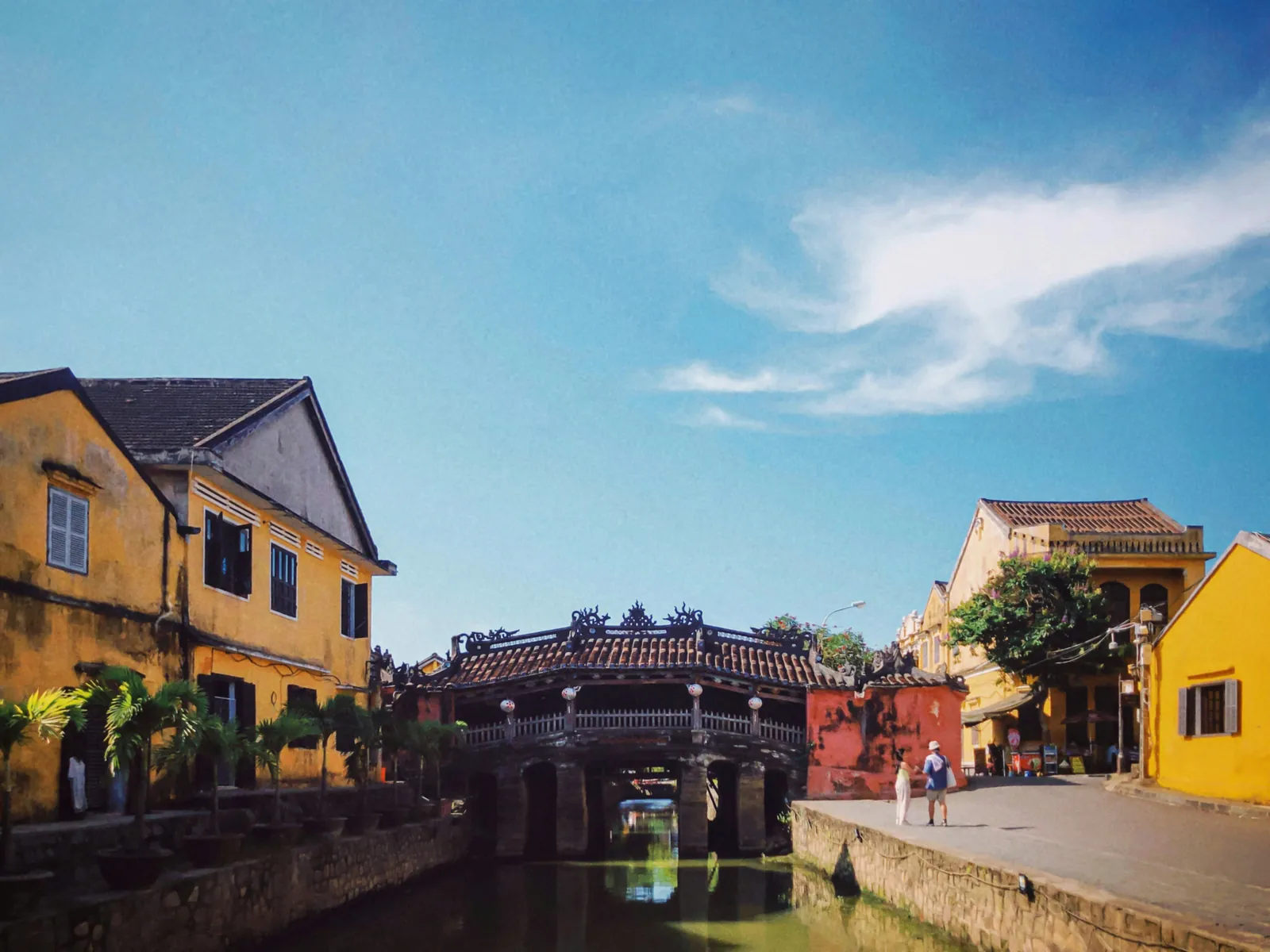 Old covered bridge with a tiled roof over a narrow canal, between ochre yellow houses under a bright blue sky